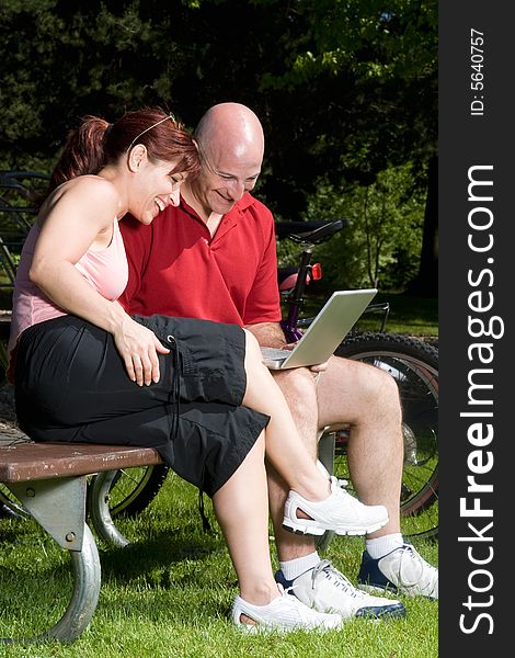 Couple on a park bench laugh as they look at a laptop computer, their bikes are in the background. Vertically framed photograph. Couple on a park bench laugh as they look at a laptop computer, their bikes are in the background. Vertically framed photograph