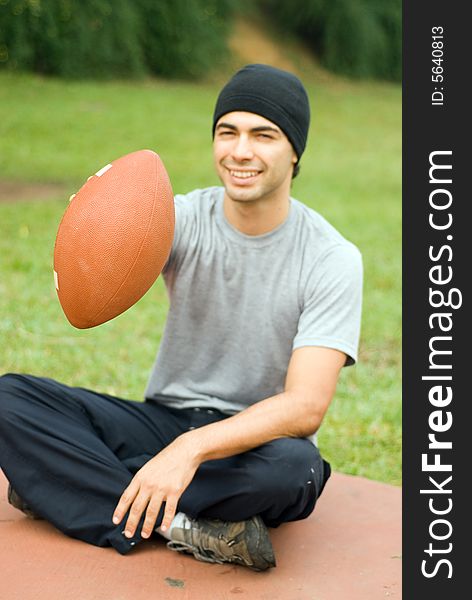 A man is sitting in a park.  He is smiling, looking at the camera and holding out a football.  Verticallly framed photo. A man is sitting in a park.  He is smiling, looking at the camera and holding out a football.  Verticallly framed photo.