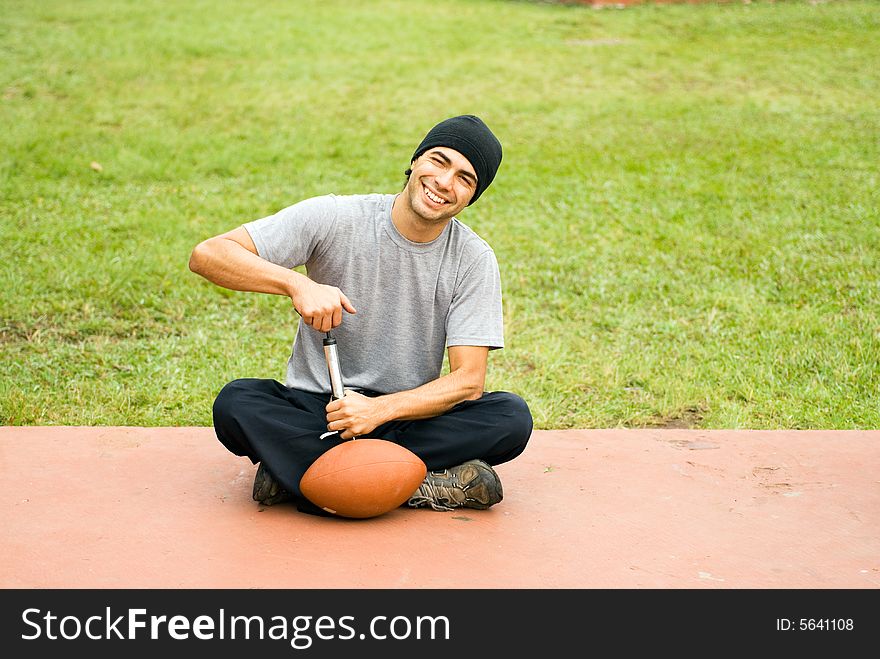A man is sitting in a park.  He is smiling, looking at the camera and pumping air into a football.  Horizontally framed photo. A man is sitting in a park.  He is smiling, looking at the camera and pumping air into a football.  Horizontally framed photo.