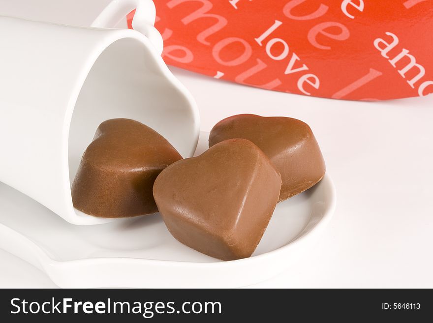 Three brown heart shaped chocolates placed on a heart shaped cup and causer with red box in background, all on white. Three brown heart shaped chocolates placed on a heart shaped cup and causer with red box in background, all on white