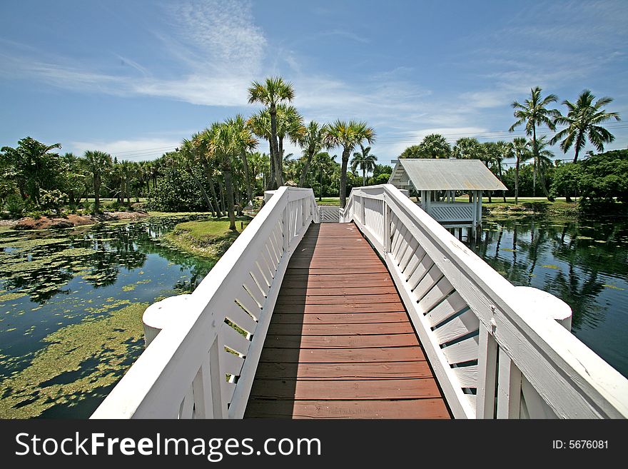 A photo of a white bridge crossing over a man made pond to an island. A photo of a white bridge crossing over a man made pond to an island.