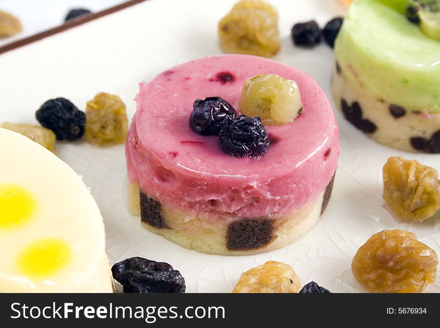 A close up of a red fruits petit four with blueberry and grape on top, surrounded by others petit fours and raisins. Main focus on the central petit four.