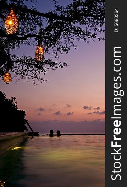 A couple takes a lantern lit dip in a cliff edge pool. Bright lanterns, silhouette tree and couple, soft sunset background. A couple takes a lantern lit dip in a cliff edge pool. Bright lanterns, silhouette tree and couple, soft sunset background.