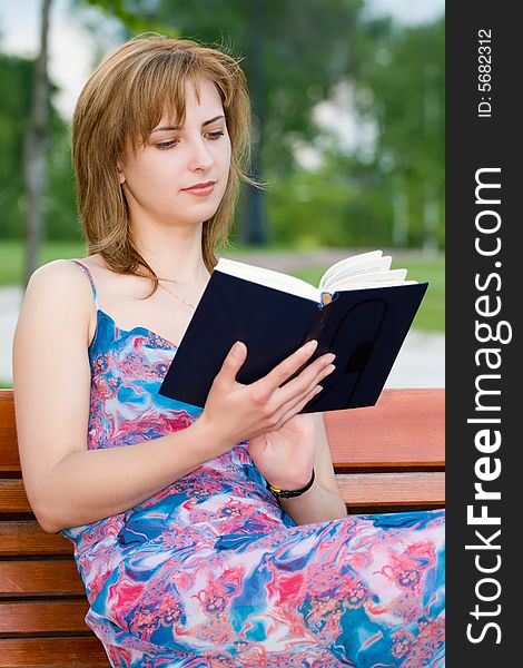 Attractive young girl reads the book sitting on a bench in park