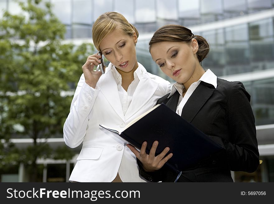 Two attractive businesswomen talking on the phone and holding a notebook outdoors. Two attractive businesswomen talking on the phone and holding a notebook outdoors