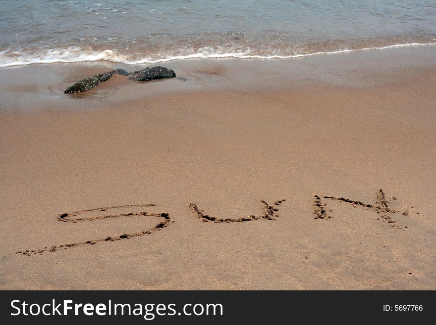 A sandy beach with the word SUN written in sand with ocean wave and seaweed in background