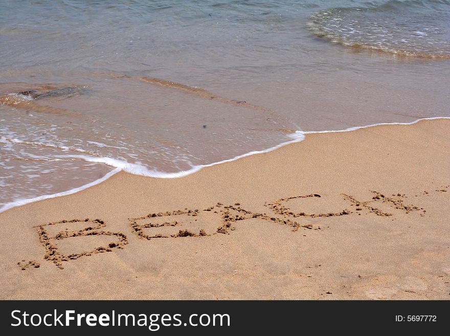 A sandy beach with the word BEACH written in sand with ocean wave and seaweed in background