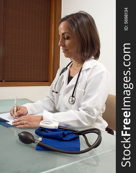 A female doctor, sits at her desk with her blood pressure tester on it, writing in her clipboard. - vertically framed. A female doctor, sits at her desk with her blood pressure tester on it, writing in her clipboard. - vertically framed