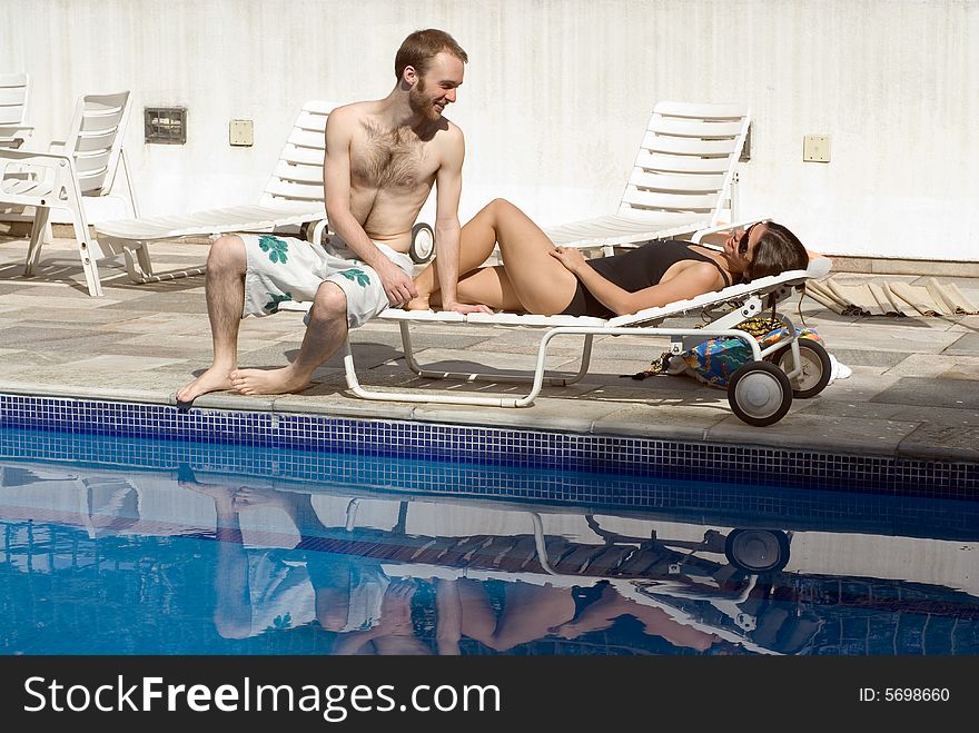 A young couple, share a pool chair, the woman lying, while the man sits near her feet. - horizontally framed. A young couple, share a pool chair, the woman lying, while the man sits near her feet. - horizontally framed