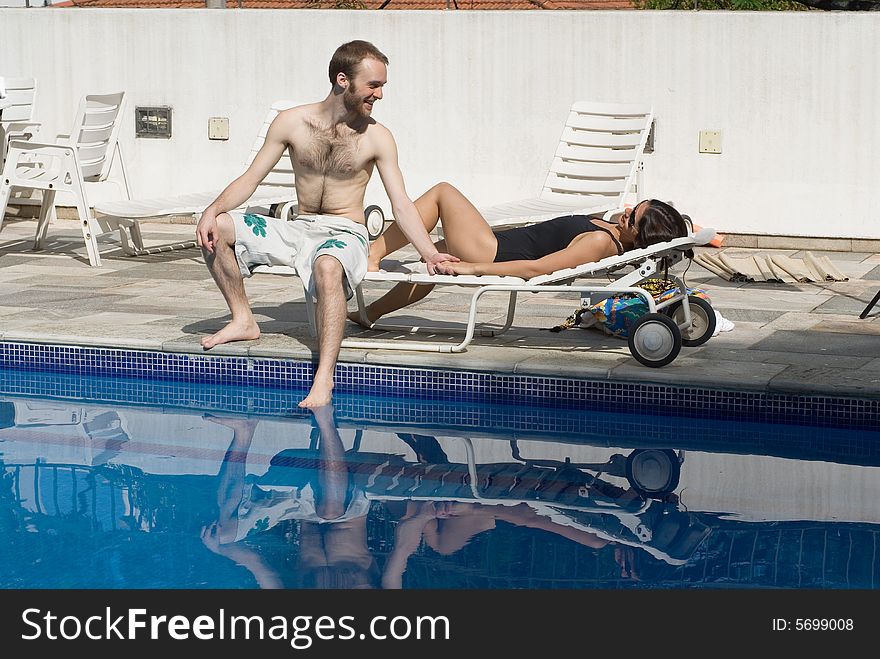 A man and a woman are relaxing next to a pool.  The man is sitting up and looking at the woman.  The woman is laying down and looking at the man.  Horizontally framed photo. A man and a woman are relaxing next to a pool.  The man is sitting up and looking at the woman.  The woman is laying down and looking at the man.  Horizontally framed photo.