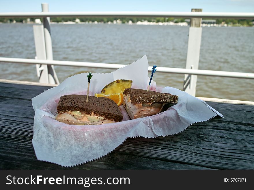 A sandwich in a basket on a rough table with a lake and shore line in the background. A sandwich in a basket on a rough table with a lake and shore line in the background
