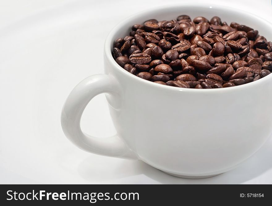 Rich brown beans fill up a classic white coffee mug sitting on large white saucer. Rich brown beans fill up a classic white coffee mug sitting on large white saucer