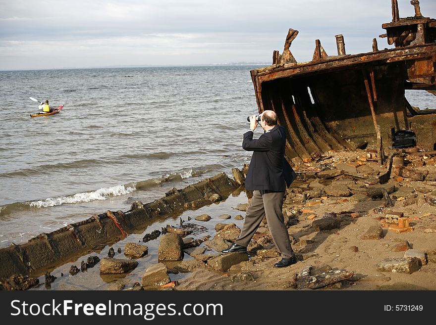 Businessman in the rusting hull of shipwreck, with binoculars looking out to the horizon where canoeist is, asking the question is their an escape from a sinking business. Businessman in the rusting hull of shipwreck, with binoculars looking out to the horizon where canoeist is, asking the question is their an escape from a sinking business