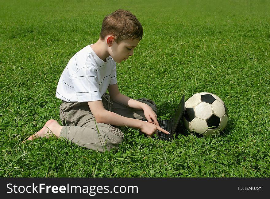The boy sitting on a grass with computer and soccer ball. The boy sitting on a grass with computer and soccer ball.