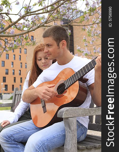 Young man plays for young girl on his guitar, it listens to him, both sits on a bank in the park. Young man plays for young girl on his guitar, it listens to him, both sits on a bank in the park