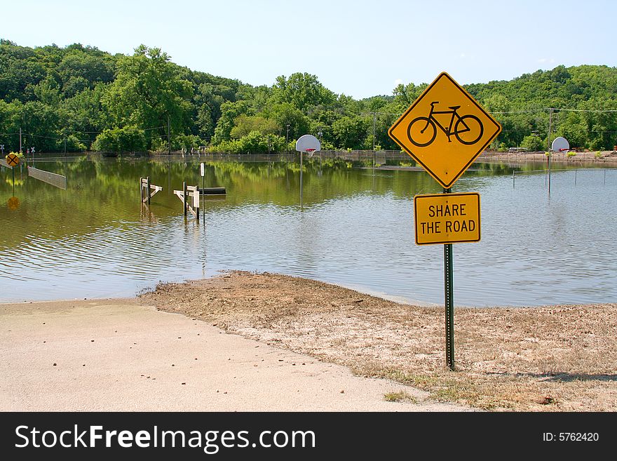 Flood waters share the road with a bicycle sign. A basketball court in the background is likewise submerged. Flood waters share the road with a bicycle sign. A basketball court in the background is likewise submerged.