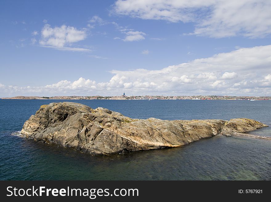 Rocky coastal island in the ocean. Blue sky with white clouds. A coastal town in the background. Rocky coastal island in the ocean. Blue sky with white clouds. A coastal town in the background.