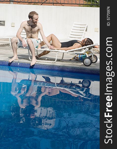 A man and a woman are relaxing next to a pool. The man is sitting up and looking at the woman. The woman is laying down and looking at the man. Vertically framed photo. A man and a woman are relaxing next to a pool. The man is sitting up and looking at the woman. The woman is laying down and looking at the man. Vertically framed photo.