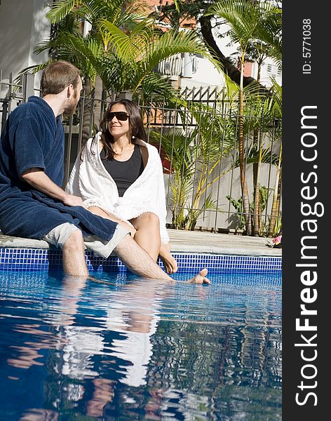 A man and a woman are relaxing together beside a pool. They are looking at each other and dipping their legs in the water. Vertically framed photo. A man and a woman are relaxing together beside a pool. They are looking at each other and dipping their legs in the water. Vertically framed photo.