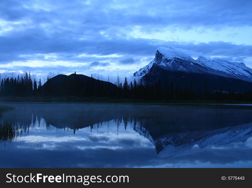 Mountains and forest reflected in a calm lake as the morning fills the sky with light. Mountains and forest reflected in a calm lake as the morning fills the sky with light