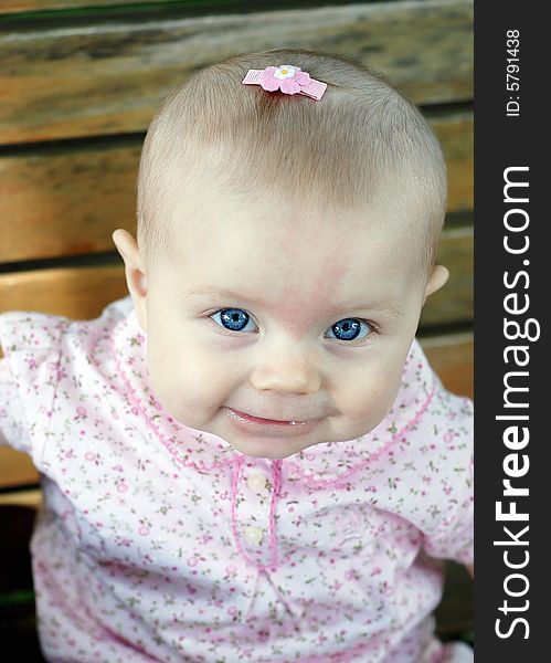 A young baby, wearing a pink bow in her hair and pink clothes, sits on a wooden park bench, smiling. Vertically framed shot. A young baby, wearing a pink bow in her hair and pink clothes, sits on a wooden park bench, smiling. Vertically framed shot.