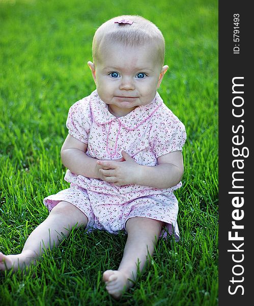 A young baby, wearing pink, sits in a grassy field, smiling. Vertically framed shot. A young baby, wearing pink, sits in a grassy field, smiling. Vertically framed shot.