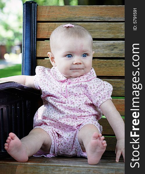 A young baby smiling while looking away, sits on a wooden park bench, wearing pink clothes. Vertically framed shot. A young baby smiling while looking away, sits on a wooden park bench, wearing pink clothes. Vertically framed shot.