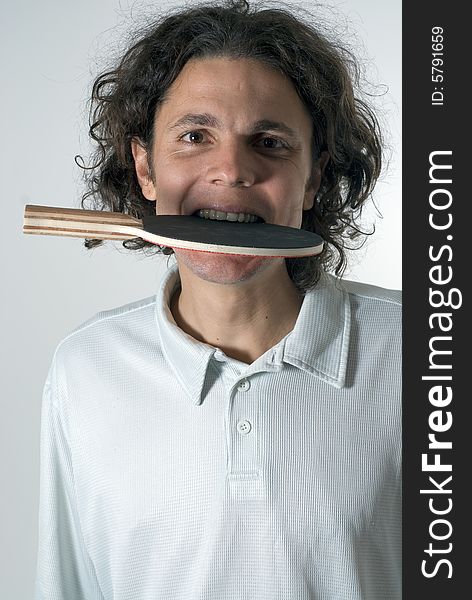 A man, standing, wearing a white polo, bites a ping pong paddle while looking at the camera. Vertically framed shot. A man, standing, wearing a white polo, bites a ping pong paddle while looking at the camera. Vertically framed shot.