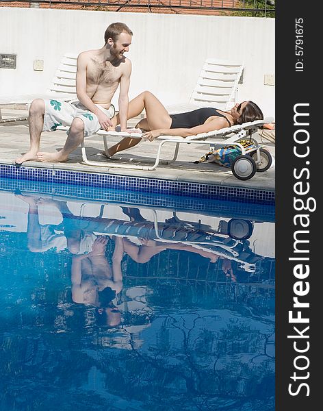 A young couple, sharing a poolside reclining chair, the woman lies down, as the man sits by her feet. - vertically framed. A young couple, sharing a poolside reclining chair, the woman lies down, as the man sits by her feet. - vertically framed