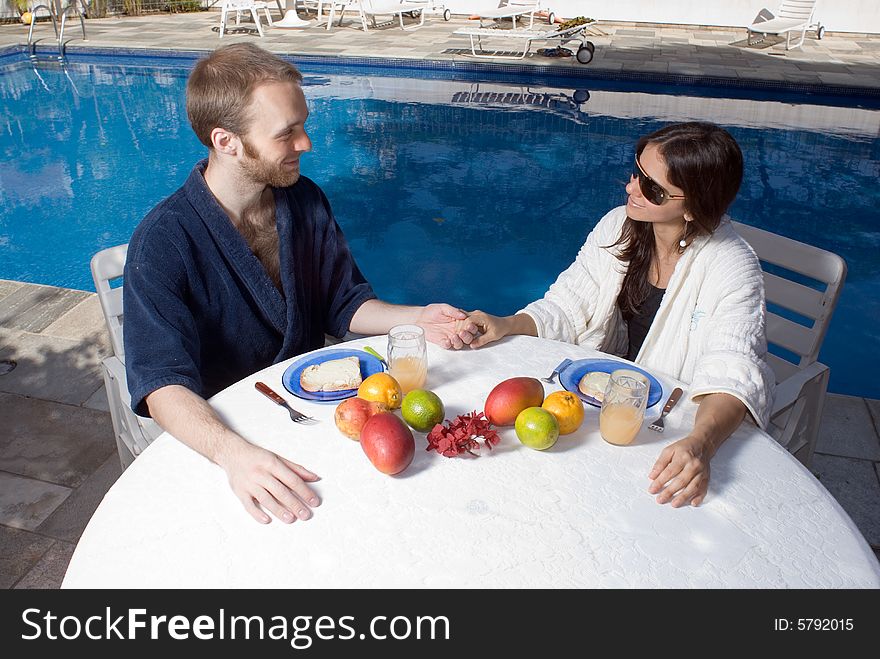 A couple, sitting at a table with fruits on it, near the pool, stare and smile, while holding hands. - horizontally framed. A couple, sitting at a table with fruits on it, near the pool, stare and smile, while holding hands. - horizontally framed