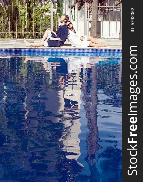 A man and a woman are relaxing together beside a pool.  They are leaning against each other.  The man is looking away from the camera and the woman is looking at it.  Vertically framed photo. A man and a woman are relaxing together beside a pool.  They are leaning against each other.  The man is looking away from the camera and the woman is looking at it.  Vertically framed photo.