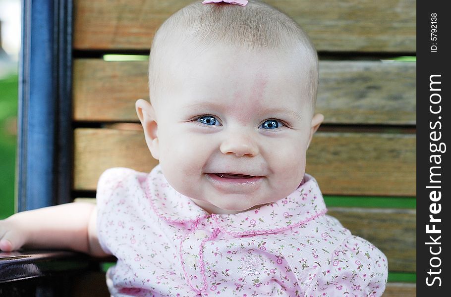 A young baby smiling while seated on a wooden park bench, wearing pink clothes. Horizontally framed shot. A young baby smiling while seated on a wooden park bench, wearing pink clothes. Horizontally framed shot.