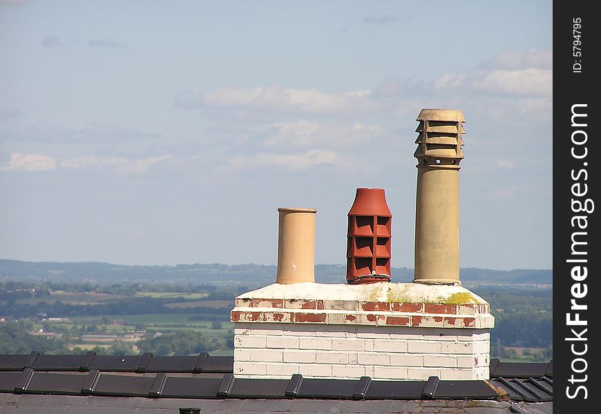 A Victorian brick chimney stack has three large chimney-pots on it.  Two are buff and the other red terracotta.  Two are louvred. Fields and woods can be seen beyond the roof.  The sky is blue with a few clouds. A Victorian brick chimney stack has three large chimney-pots on it.  Two are buff and the other red terracotta.  Two are louvred. Fields and woods can be seen beyond the roof.  The sky is blue with a few clouds.