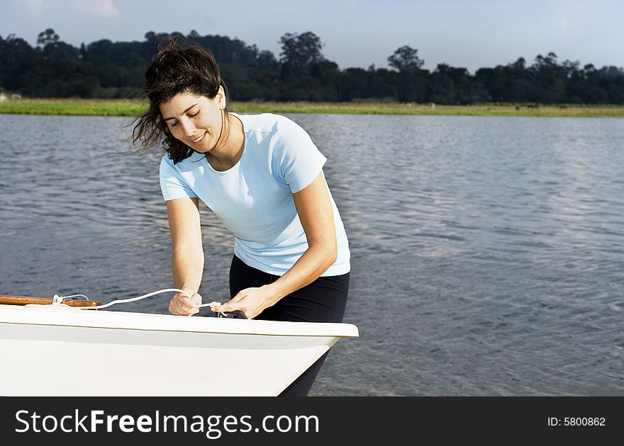 A woman is standing next to a sailboat on the water.  She is tying a knot in a rope, smiling, and looking away from the camera.  Horizontally framed shot. A woman is standing next to a sailboat on the water.  She is tying a knot in a rope, smiling, and looking away from the camera.  Horizontally framed shot.