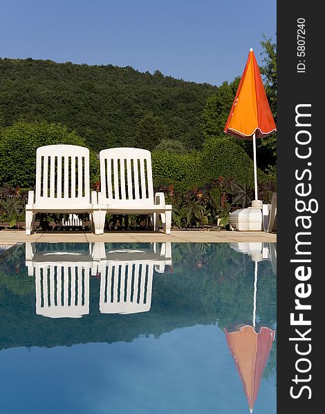 Two white sunloungers reflected in a blue swimming pool, next to an orange umbrella. Two white sunloungers reflected in a blue swimming pool, next to an orange umbrella