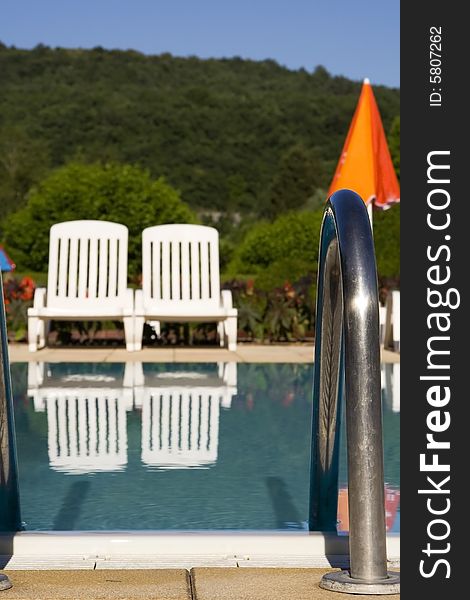 Two white sunloungers reflected in a blue swimming pool, next to an orange umbrella.  The focus is on the metal handrail in the foreground. Two white sunloungers reflected in a blue swimming pool, next to an orange umbrella.  The focus is on the metal handrail in the foreground.