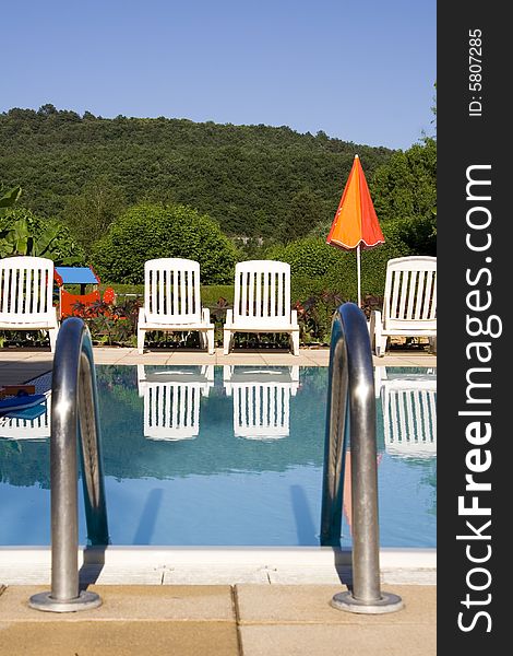 White sunloungers reflected in a blue swimming pool, next to an orange umbrella. White sunloungers reflected in a blue swimming pool, next to an orange umbrella