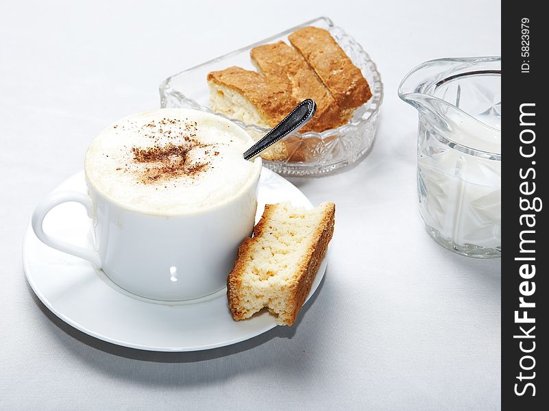 Coffee in white cup with spoon and caramal rusk, more rusks and milk in background. Coffee in white cup with spoon and caramal rusk, more rusks and milk in background