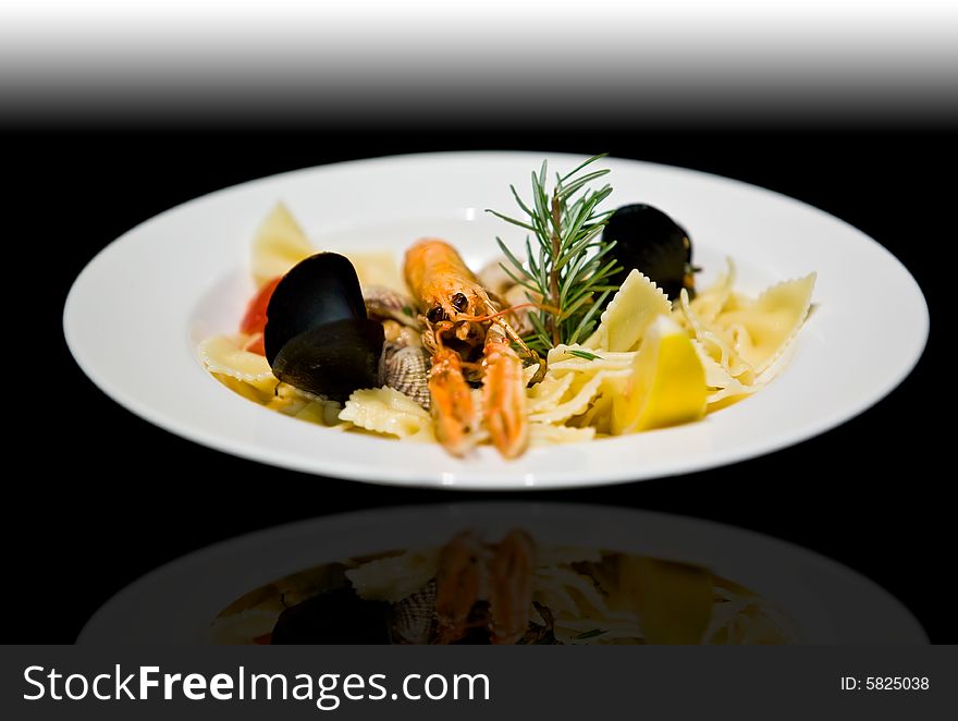 Langoustine, clam, and mussel pasta appetizer served with rosemary and lemon in a white plate over a graduate black background. Langoustine, clam, and mussel pasta appetizer served with rosemary and lemon in a white plate over a graduate black background.