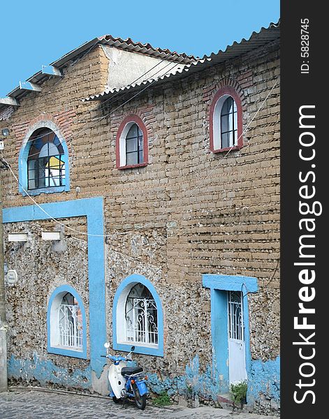 Blue motorbike parked alongside a stone building accented in blue trim, image of a poor Mexican village.