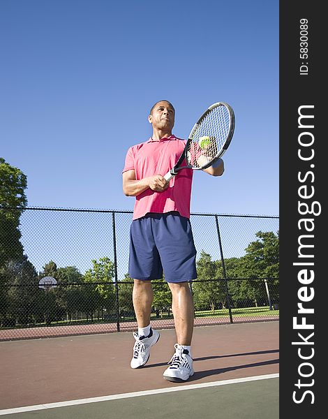 A man is outside on a tennis court playing tennis. He is about to serve the ball and is looking away from the camera. A man is outside on a tennis court playing tennis. He is about to serve the ball and is looking away from the camera.