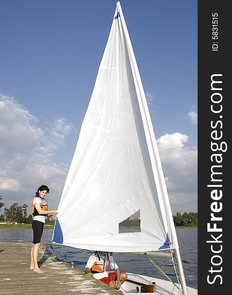 A woman and man are next to a sailboat.  The man is sitting inside the boat.  The woman is standing beside it fixing the sail and smiling at the camera.  Vertically framed shot. A woman and man are next to a sailboat.  The man is sitting inside the boat.  The woman is standing beside it fixing the sail and smiling at the camera.  Vertically framed shot.