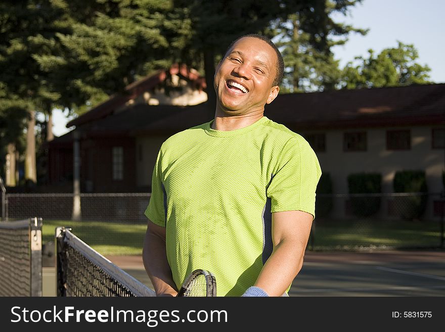 A man is outside on a tennis court.  He is leaning against the net and smiling at the camera. A man is outside on a tennis court.  He is leaning against the net and smiling at the camera.