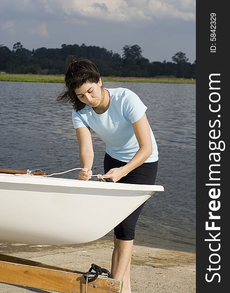 A woman is standing next to a sailboat on a lake.  She is tying a knot and looking at the rope.  Vertically framed photo. A woman is standing next to a sailboat on a lake.  She is tying a knot and looking at the rope.  Vertically framed photo.