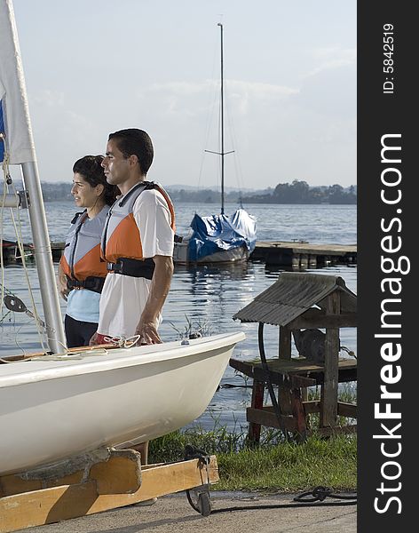 A young couple is standing next to a sailboat.  They are smiling and looking away from the camera.  Vertically framed shot. A young couple is standing next to a sailboat.  They are smiling and looking away from the camera.  Vertically framed shot.