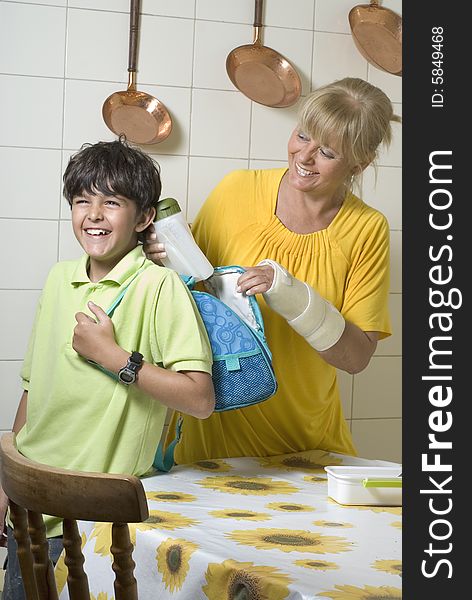 Woman standing behind boy filling backpack in kitchen. Woman smiling at boy. Vertically framed photo. Woman standing behind boy filling backpack in kitchen. Woman smiling at boy. Vertically framed photo.