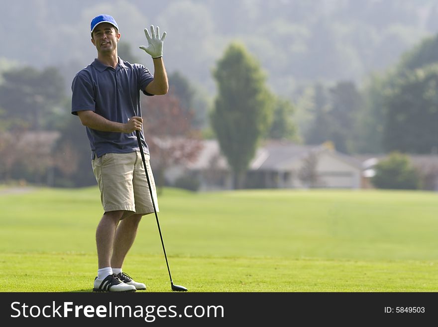 Man standing on golf course waving his right hand at the camera with left hand resting on golf club - horizontally framed photo. Man standing on golf course waving his right hand at the camera with left hand resting on golf club - horizontally framed photo