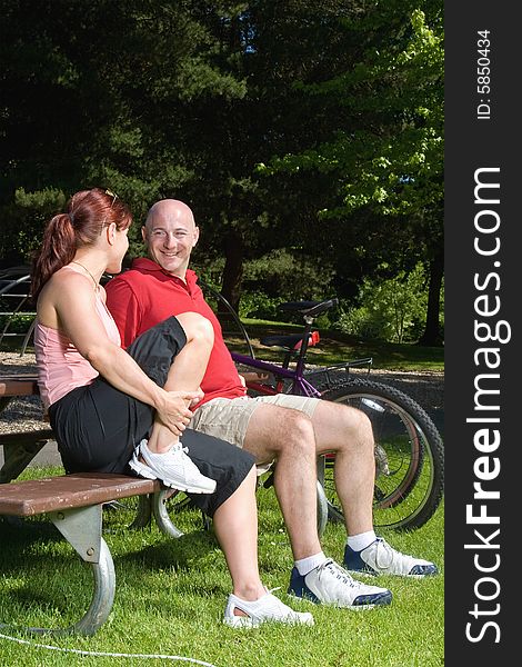 Happy couple sitting on a park bench next to their bicycles. They are looking at each other and smiling. Vertically framed photograph. Happy couple sitting on a park bench next to their bicycles. They are looking at each other and smiling. Vertically framed photograph