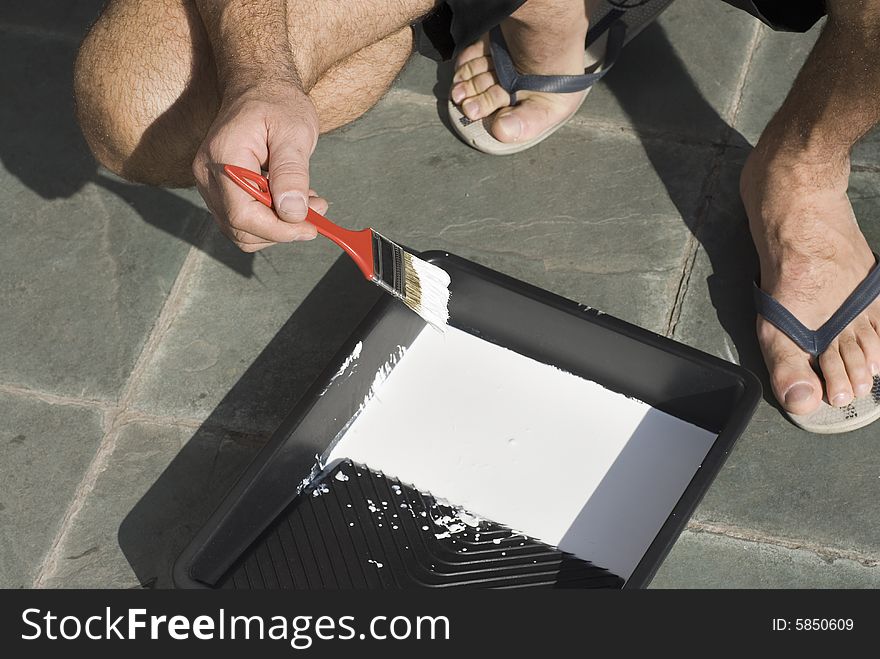 A man, kneels, dipping his paintbrush into a platter of white paint. Horizontally framed shot. A man, kneels, dipping his paintbrush into a platter of white paint. Horizontally framed shot.