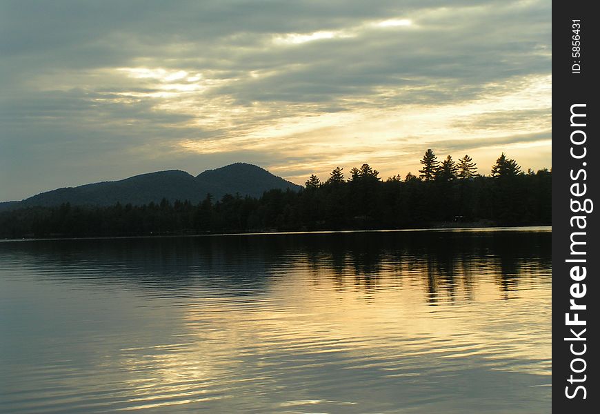 Mountain and trees silhouetted at sunset with clouds reflecting on lake in Adirondacks. Mountain and trees silhouetted at sunset with clouds reflecting on lake in Adirondacks.